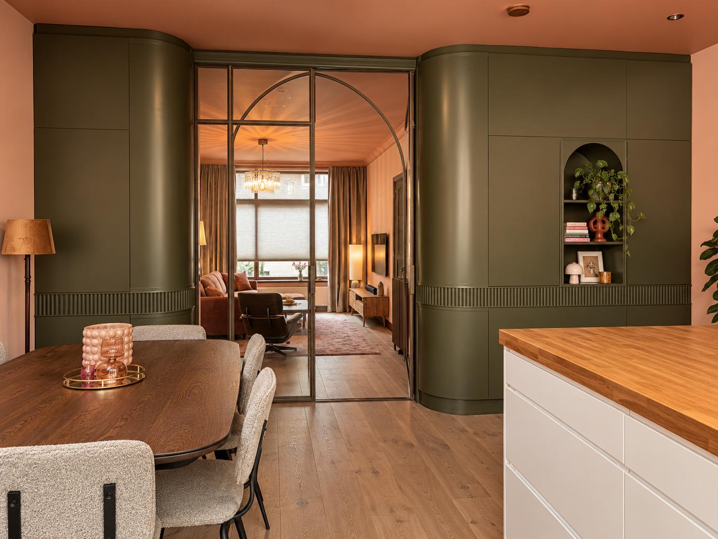 Keuken met olijfgroen maatwerk, terracotta plafond en een stalen taatsdeur met boog naar de woonkamer